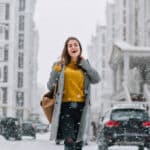 Outdoor photo of graceful female model in trendy long coat relaxing in city in winter day. Good looking young woman in yellow sweater having fun during shopping in december morning..